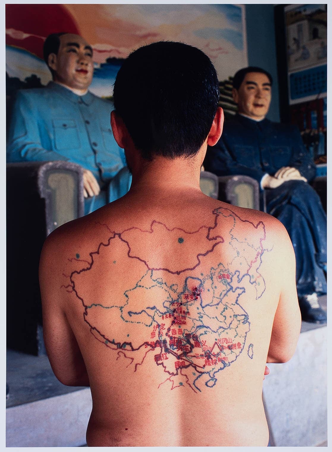 A man with a tattoo of a map of China on his back stands in front of statues of Mao Zedong and another leader. The image blends themes of identity, geography, and political history.