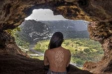 Topless medium-skinned person with a Mesoamerican-style drawing or tattoo on their back looking out a hole in a rock formation at a green field in the distance