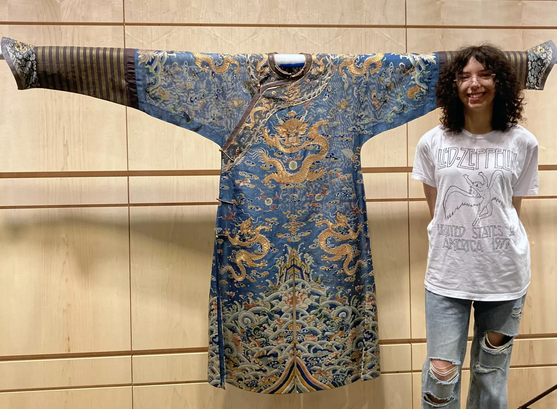 A person standing next to a blue and gold traditional Chinese dragon robe, which is richly embroidered with dragon and cloud motifs, displayed on a wall. The person is smiling and wearing a Led Zeppelin t-shirt and ripped jeans.