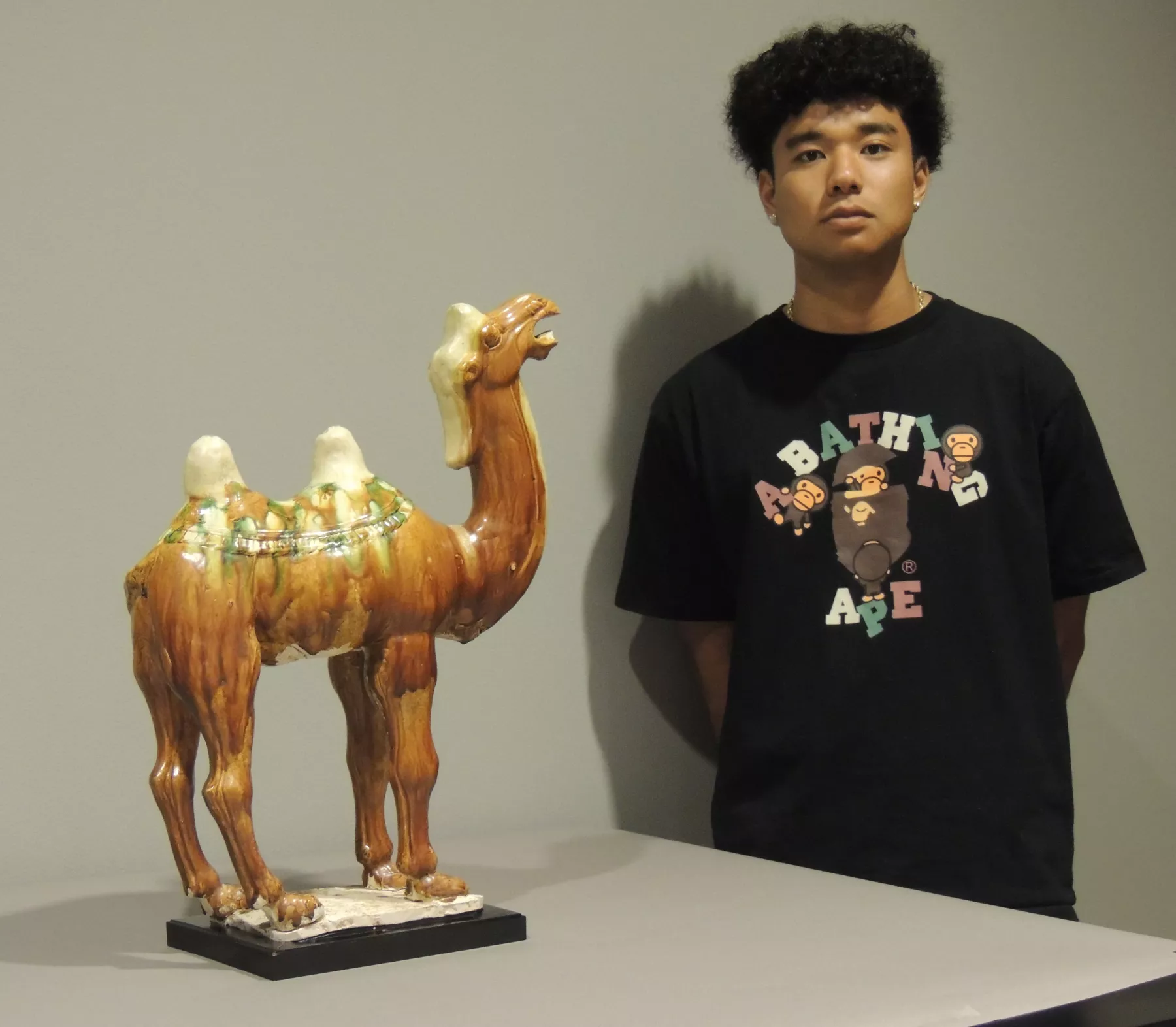 A young man standing next to a ceramic camel. He has a black t-shirt with the words "A Bathing Ape" on it with images of monkeys.